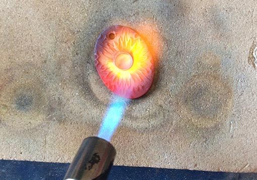 A handheld torch with a flame that is heating a piece of metal clay to fire it.